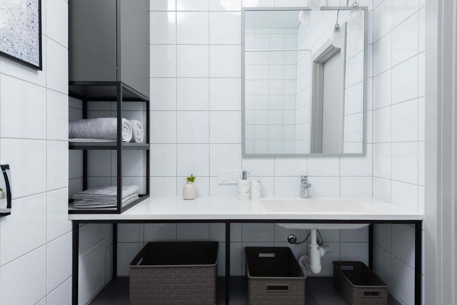 Interior of light bathroom with white counter near shelves with containers and towels near sink with faucet and mirror on tile near soap dispenser