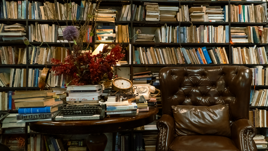 A vintage study room featuring a leather armchair, bookshelves, and a rustic table with decor.