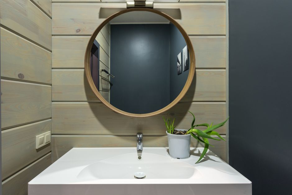 Round mirror hanging above sink with faucet and potted plant on wall made of panels in modern bathroom